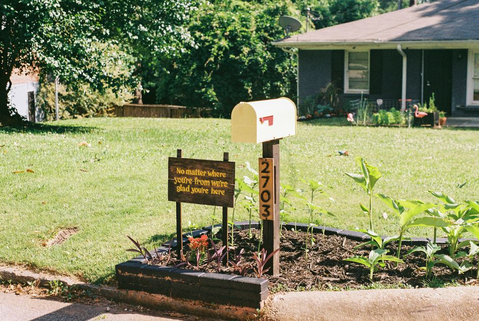 A small front yard with a neatly maintained flower bed bordered by dark bricks, containing young green plants and a few purple-leaved plants. A wooden sign with yellow text reading 'No matter where yo